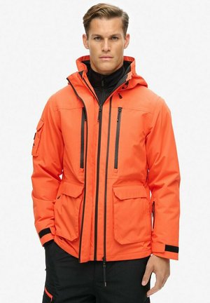 Orange vandtæt jakke med hætte, der har sorte lynlåse, to brystlommer og justerbare manchetter. Teksturert stof for holdbarhed.