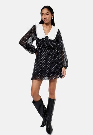 Vrouw met een zwarte doorschijnende polkadotjurk met een grote witte kraag, lange pofmouwen en zwarte kniehoge laarzen, staand met één been gekruist.