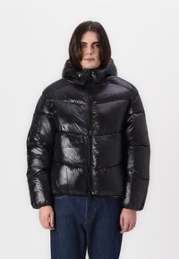 Sorte pufferjakke med en blank finish, der har høj krave, hellynlås og quiltet design. Letvægts og isoleret for varme.