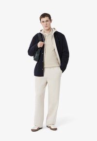 Beige hoodie under a mörk marinblå jacka, tillsammans med ljusbeiga byxor och vita skor med bruna accenter. Inkluderar en liten grön väska.