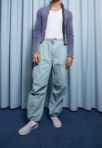 Ljusblå cargobyxor med elastiska muddar, en formad vit linne och en ribbad lila zip-up kofta. Matchas med lila rutiga slip-on skor.