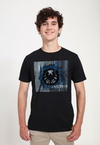 Černé bavlněné tričko s grafikou stylizovaného orlího loga v modré a šedé barvě na texturovaném pozadí, orámované v obdélníkovém designu.