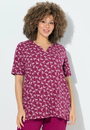 Donna con capelli ricci che indossa orecchini a cerchio, una maglietta bordeaux a maniche corte con motivo scritto bianco e pantaloni bordeaux abbinati, in piedi contro un muro bianco.