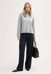 Pull en tricot gris avec un col montant, associé à un pantalon large bleu marine. Le mannequin tient un sac à main en cuir noir. Textures lisses, design minimaliste.