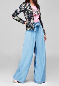 Blomsterprint bomberjakke, sort med pink og grønne accenter; lys blå brede denimbukser; modellen har en pink t-shirt med grafisk tryk på under.