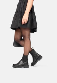 Stivaletti neri in pelle con suola spessa, pannelli laterali elasticizzati e una zip sul retro. Indossati con un vestito nero e collant.