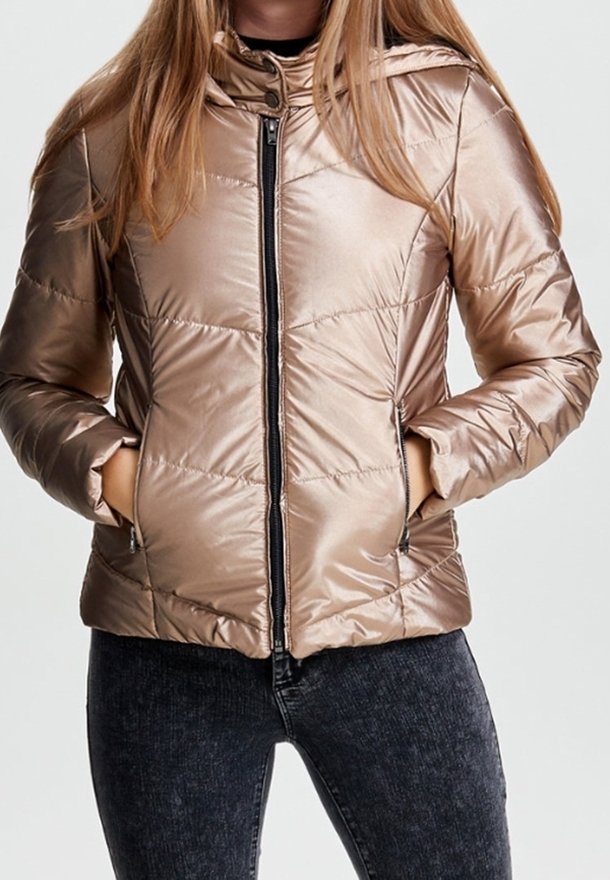 Vrouw in een glanzende beige pufferjack met handen in de ritszakken, gecombineerd met donker gewassen jeans.