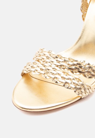 Χρυσό σανδάλι ανοιχτού δακτύλου με πλεγμένα μεταλλικά λουριά μπροστά και λείο, γυαλιστερό πέλμα σε απλό λευκό φόντο.