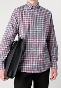 Mann in kariertem Hemd mit roten, blauen und weißen Farben, der eine schwarze Ledertasche seitlich hält und schwarze Hosen trägt.