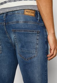 Jeans de denim azul ajustados con un acabado texturizado, que presentan una etiqueta de cuero marrón y dos bolsillos traseros con detalles de costura visibles.