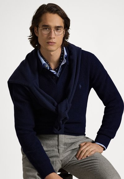Marineblå strikket genser med sjalskrage, kombinert med en lyseblå skjorte med hvite prikker og grå teksturerte bukser.