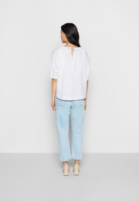 Blusa branca com mangas bufantes e um detalhe de abertura nas costas, combinada com jeans azul claro e sapatos de salto bloco beges. Textura de tecido suave.