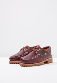 Bateau en cuir bourgogne foncé avec un design en surpiqûres surélevées, lacets marron, œillets en métal et semelle en caoutchouc texturée.