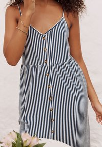 Stribet kjole med mørkeblå og hvide lodrette linjer, V-udskæring, justerbare stropper, knaplukning foran og indsnævret talje med en flydende nederdel.