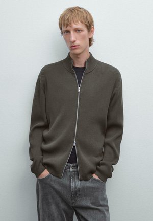 Young man with blonde hair wearing a dark olive ribbed zip-up sweater over a black shirt, paired with faded gray jeans, hands in pockets.