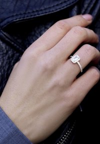 Anello d'argento con una pietra preziosa trasparente rettangolare, circondata da piccoli diamanti, su una mano umana su uno sfondo nero texturizzato.