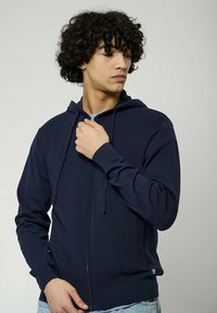 Sweat à capuche zippé bleu marine en tissu texturé, avec un cordon de serrage à la capuche et des poignets côtelés. Présente un logo discret sur l'ourlet et une coupe ajustée.