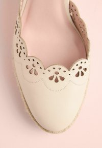 Beige espadrille with scalloped edge, perforated floral design, leather upper, jute sole, and smooth texture. Pink background.