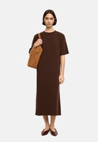 Brąsowa, dzianinowa sukienka do kolan z krótkimi rękawami. Modelka trzyma beżową, zamszową torebkę na ramię i nosi ciemnoczerwone płaskie buty. Prosty design, bez wzorów.