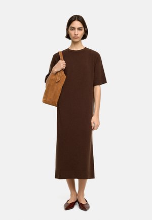 Brąsowa, dzianinowa sukienka do kolan z krótkimi rękawami. Modelka trzyma beżową, zamszową torebkę na ramię i nosi ciemnoczerwone płaskie buty. Prosty design, bez wzorów.
