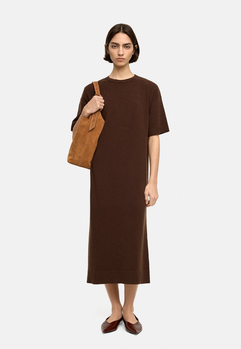 Brąsowa, dzianinowa sukienka do kolan z krótkimi rękawami. Modelka trzyma beżową, zamszową torebkę na ramię i nosi ciemnoczerwone płaskie buty. Prosty design, bez wzorów.