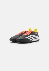 Zwarte sportschoenen met een gestructureerde bovenkant, witte strepen, feloranje veters en een gele binnenvoering. Ontworpen voor kunstgras, met rubberen noppen.