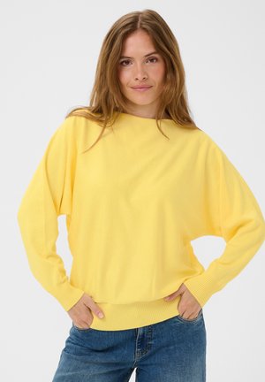 Donna con capelli lunghi castano chiaro che indossa un maglione giallo vivace e jeans blu, in piedi con le mani nelle tasche su sfondo bianco.