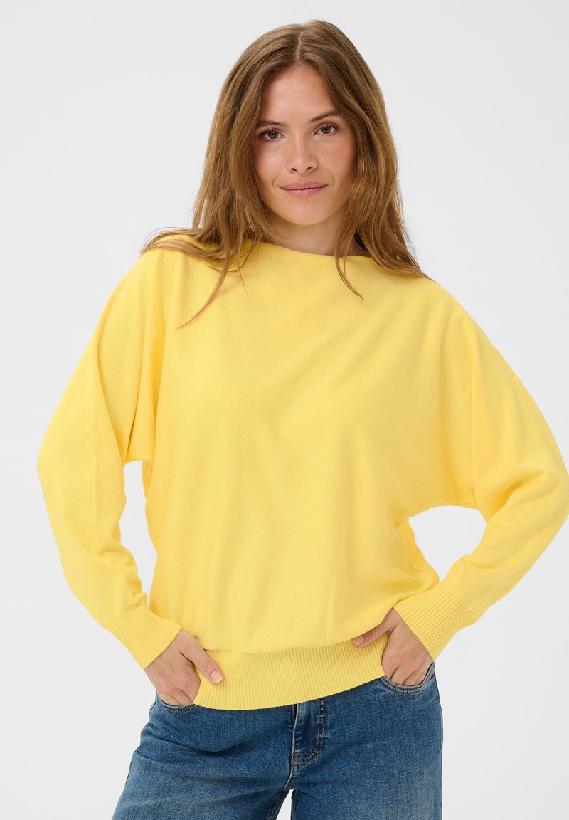 Femme aux longs cheveux châtain clair portant un pull jaune vif et un jean bleu, debout les mains dans les poches devant un fond blanc.