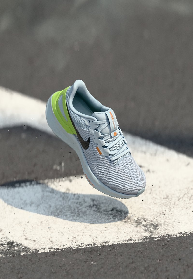 Chaussure de course Nike gris clair avec un détail vert et un swoosh noir, en l'air au-dessus d'un bitume avec une marquage blanc au sol et du gravier éparpillé.