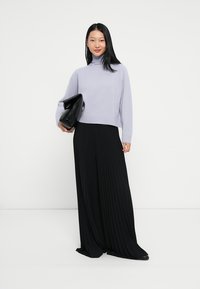 Lila Rollkragenpullover mit langen Ärmeln, kombiniert mit einem schwarzen, plissierten Maxikleid. Das Model hält eine schwarze Clutch.