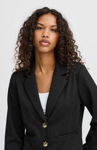 Schwarzer Blazer aus glattem Stoff, mit Reverskragen, zwei vorderen Taschen und zwei hellen Knöpfen. Das Model trägt ein weißes Oberteil.