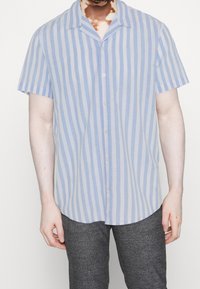 Mężczyzna w krótkim rękawie, jasnoniebieskiej i białej pionowo prążkowanej koszuli zapinanej na guziki oraz ciemnoszarych spodni w kratę, stojący z rękami w luzie.