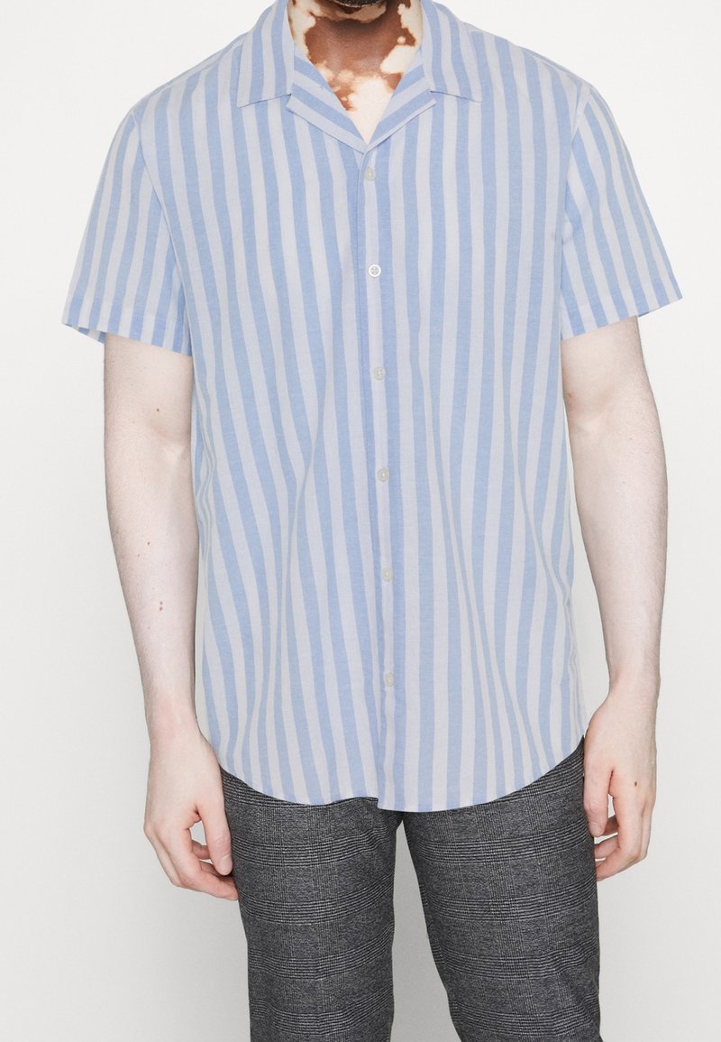 Mężczyzna w krótkim rękawie, jasnoniebieskiej i białej pionowo prążkowanej koszuli zapinanej na guziki oraz ciemnoszarych spodni w kratę, stojący z rękami w luzie.