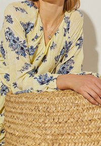 Blusa floral amarilla con patrones azules, tela texturizada y mangas con fruncidos. Una cesta de mimbre en el primer plano añade una textura natural.
