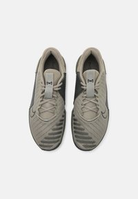Lys beige og mørkegrønne træningssko med mesh-overdel, teksturerede sider, grå snørebånd og et logodetalje på siden. Flad gummisål.