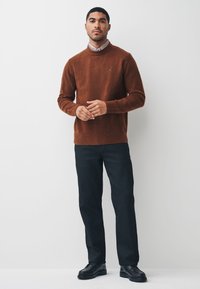 Strukturierter brauner Strickpullover mit hohem Kragen, kombiniert mit dunkler Hose und schwarzen Schuhen, mit kleinem Logo auf der Brust.