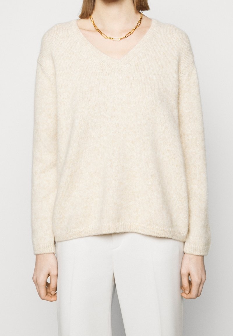 Personne portant un pull crème col V, un pantalon blanc et un gros collier chaîne en or, debout devant un fond clair uni.