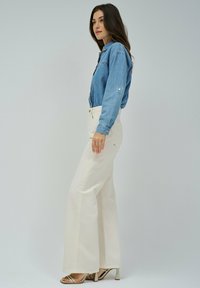 Camisa de mezclilla azul claro con cierre de botones, combinada con pantalones blancos de pierna ancha y cintura alta. La modelo lleva sandalias de tacón plateadas.