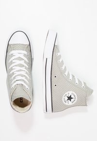 Hoge sneakers in lichtgrijs mesh met witte rubberen accenten, voorzien van witte veters en een cirkelvormige logo patch aan de buitenkant.