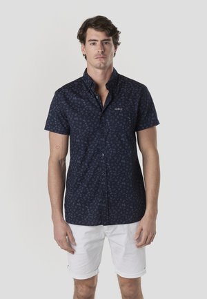 Cămașă bleumarin cu mâneci scurte, cu nasturi și un model subtil de icoane. Țesătura pare ușoară, cu o textură netedă. Pantaloni scurți albi purtați dedesubt.