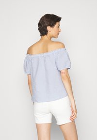 Blusa à off-the-shoulder em riscas azuis e brancas com decote elástico e mangas bufantes, combinada com calções brancos. Textura de tecido suave.