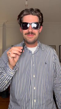 Homem usando óculos escuros e uma camisa de botão listrada. Segura um microfone preto pequeno, tem bigode e cabelo curto.