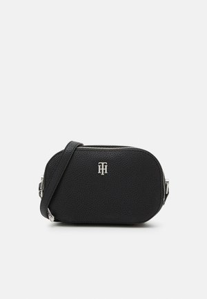 Sac en bandoulière noir texturé avec une fermeture éclair en argent et le logo "TH" sur le devant, présentant une sangle ajustable sur un fond blanc.