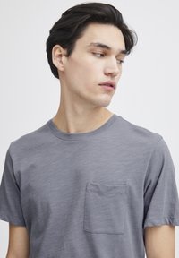 Tricou gri cu guler rotund, confecționat dintr-un material moale, având mâneci scurte și un buzunar pe piept cu o textură subtilă.