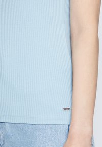 Light blue ribbed t-shirt with a round neckline, featuring a small silver logo tag at the hem, paired with light denim shorts.