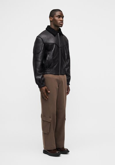 Άνδρας στέκεται, φοράει μαύρο δερμάτινο μπουφάν με γιακά από shearling, καφέ παντελόνι cargo και σκούρα καφέ παπούτσια, μπροστά από ένα απλό φωτεινό φόντο.