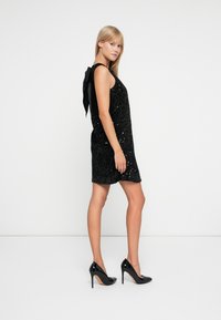Mini abito di paillettes nere con design senza maniche, caratterizzato da un grande fiocco nero sul retro e abbinato a tacchi alti neri lucidi.