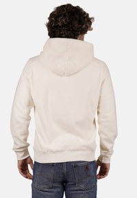 Hoodie di colore crema con un tessuto a trama e maniche aderenti, dotato di cappuccio con coulisse e una vestibilità rilassata, abbinato a jeans blu.