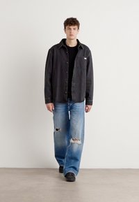 Black button-up shirt made of cotton, paired with light blue distressed jeans featuring rips. Worn with black shoes, model stands on a neutral background.
