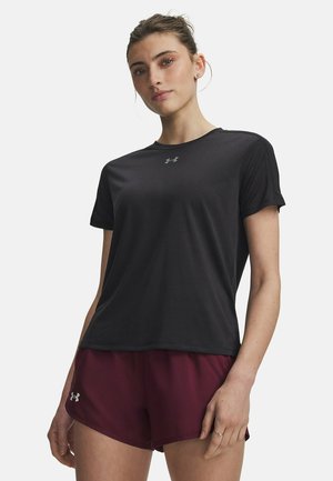 Jonge vrouw met donker haar in een knot, gekleed in een zwart sportshirt met korte mouwen en bordeauxrode sportshorts tegen een effen achtergrond.